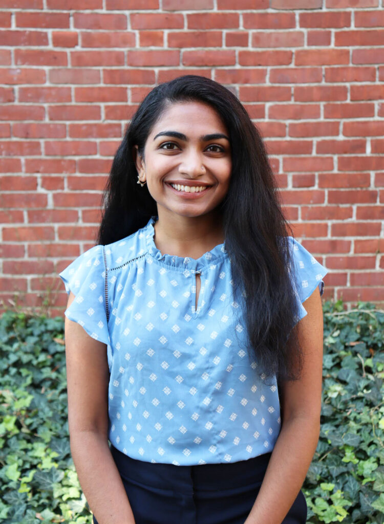 Anu Joy short-sleeved light blue shirt with white polka dots, standing against brick wall
