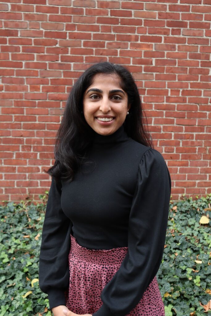 M. Patel headshot, in a black sweater standing in front of a brick wall.