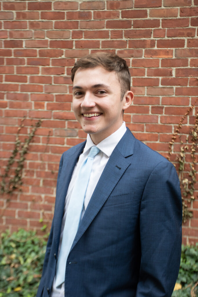 H. Patterson headshot-blue suit jacket, white shirt, in front of brick wall.