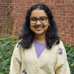 K. Sengupta headshot with white cardigan, purple shirt, in front of brick wall.