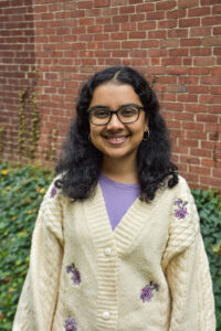 K. Sengupta headshot with white cardigan, purple shirt, in front of brick wall.