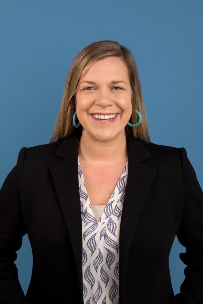Headshot Betsy Fordyce wearing a gray/white blouse, black blazer, with blue background.