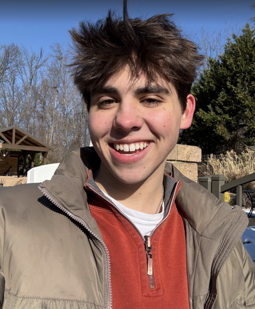 Headshot C. Colvin, brown jacket, orange shirt, trees and sky in the background.