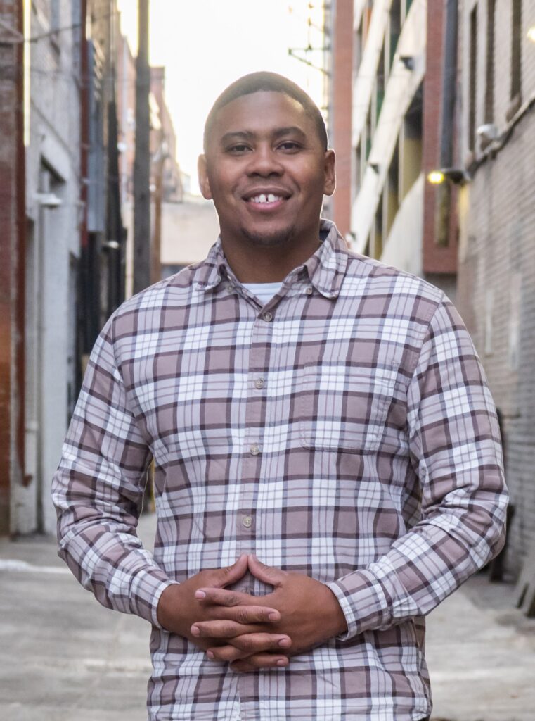 J. Henderson headshot, wearing plaid shirt, standing outdoors in front of buildings.