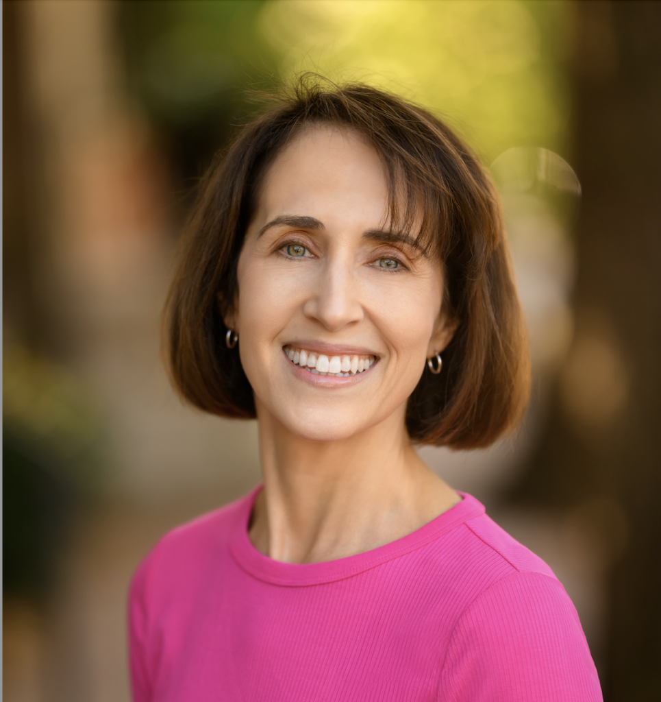 R. Belin headshot, pink sweater, nature blurred background.