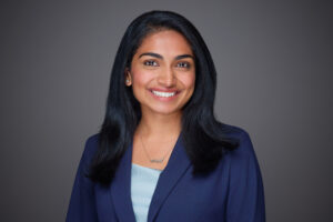 Anu Joy wearing a navy blazer, light blue blouse, and standing in front of a gray background.