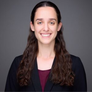Haylee Levin wearing a black blazer and burgundy blouse, standing in front of a dark gray background.
