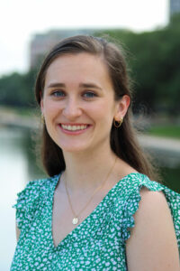 Sarah Berton wearing a teal and white blouse, standing in front of landscape with water.