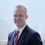 Bobby Stroup in a navy suit, red and gold tie, white shirt, standing in front of a window.