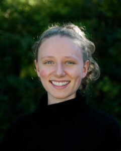 Headshot Anna Hackel in black sweater, in front of leafy background.