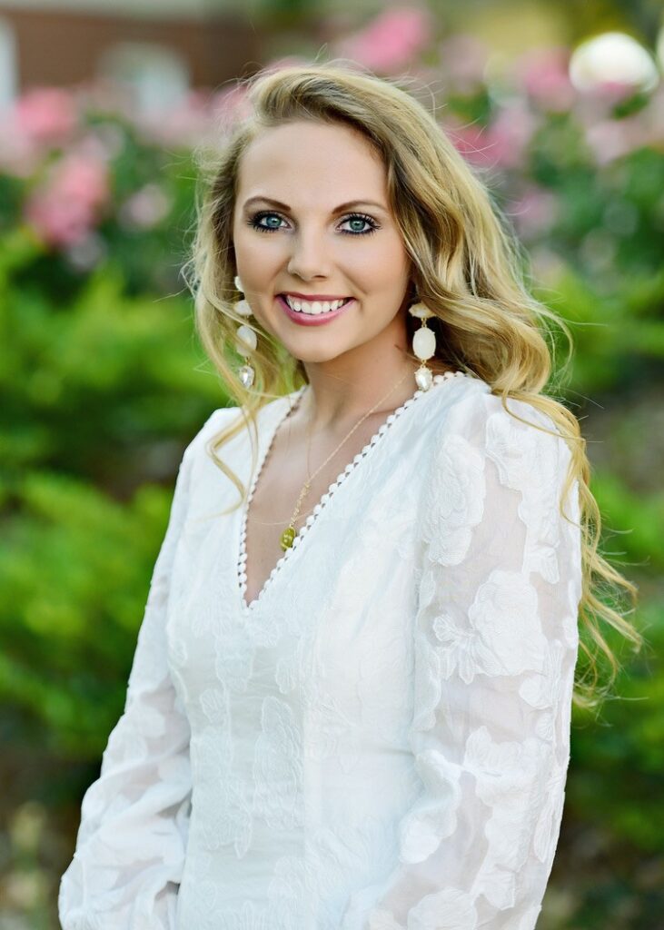 Jenny Herrell headshot, white dress standing in front of leafy background.
