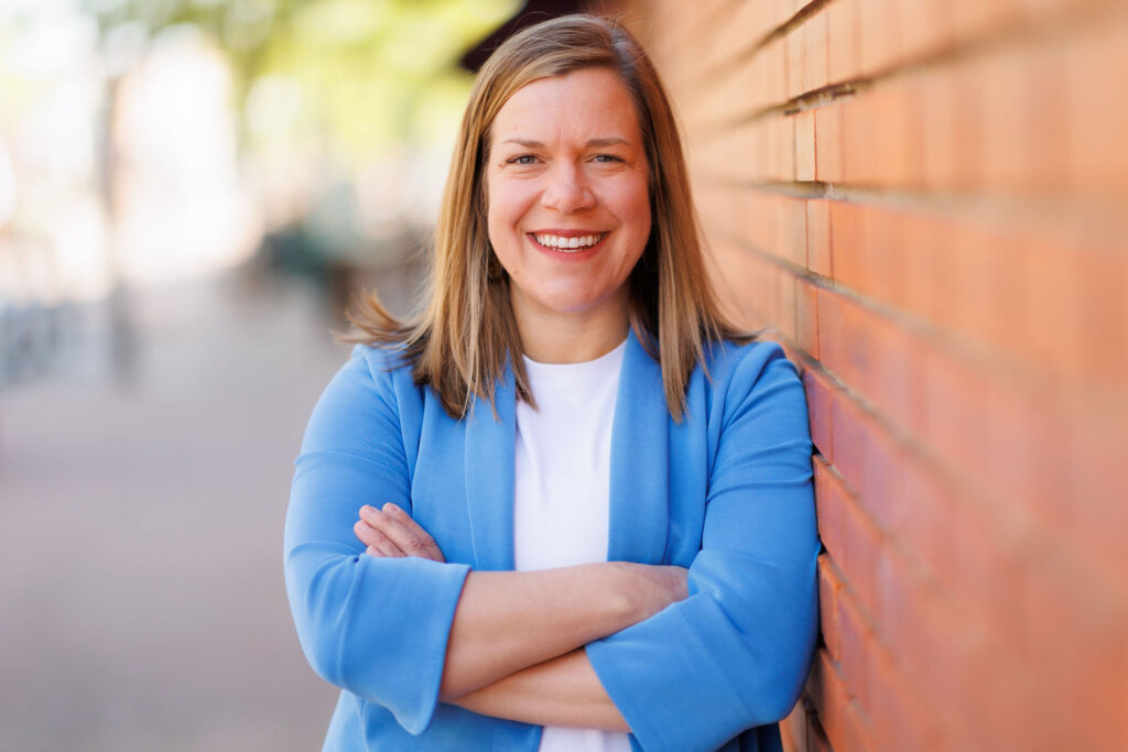Headshot of Betsy Fordyce in periwinkle jacket, leaning against brick wall.