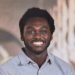 Cordy McJunkins headshot, gray shirt standing in front of white campus buildings.