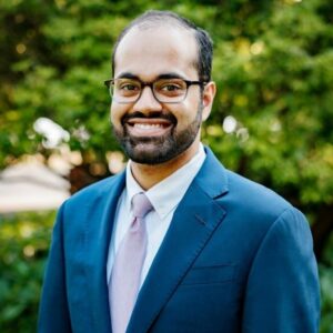 Headshot Omar Tariq, navy blazer, white shirt, lavender tie, standing in front of foliage.