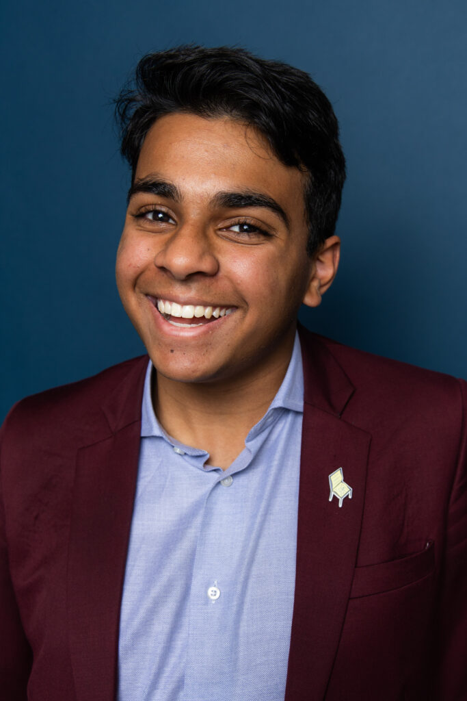 Ayaan Moledina headshot, burgundy blazer and light blue shirt, in front of dark blue background.