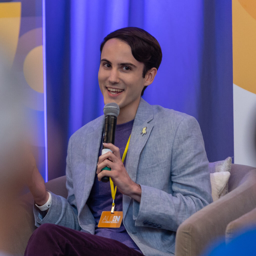 Cameron Samuels headshot, gray blazer, dark blue shirt & seated in front of blue curtains.