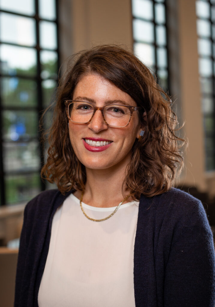 Daniela DiGiacomo headshot, standing in front of windows, wearing dark sweater with white top.