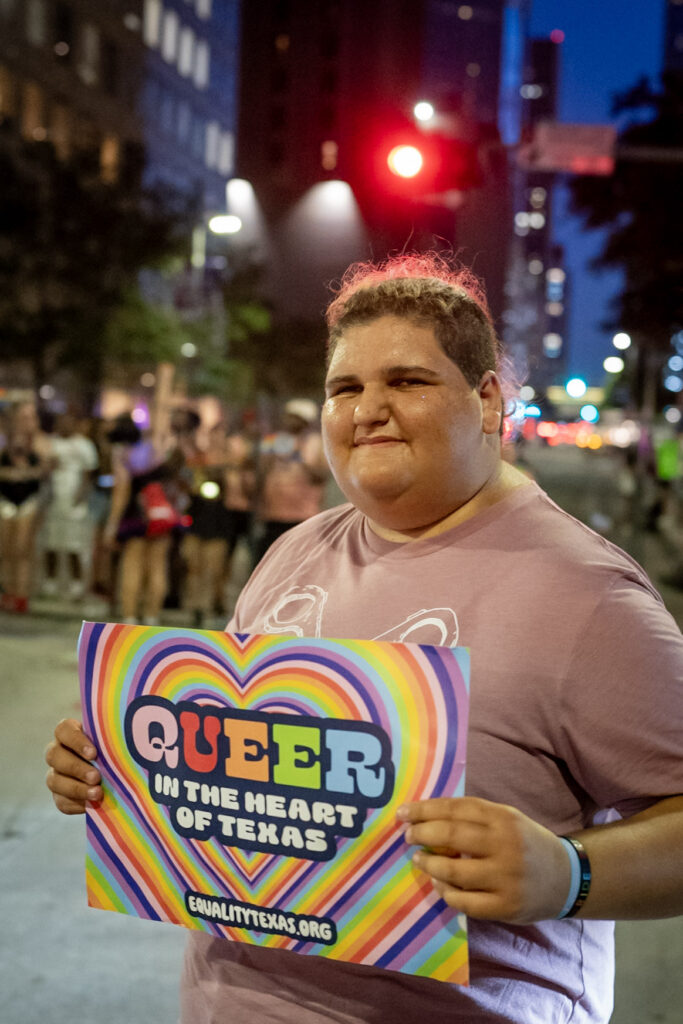 Hayden Cohen standing in front of city lights, holding rainbow color sign in a pink tee-shirt.