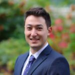 Sam Morimoto headshot in dark blue suit, dark blue tie, white shirt, standing in front of flowers and plants.