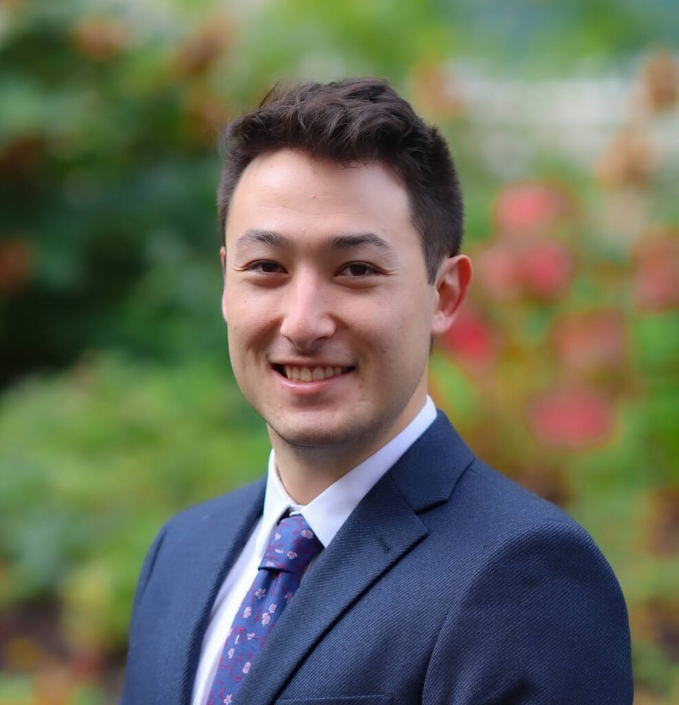 Sam Morimoto headshot in dark blue suit, dark blue tie, white shirt, standing in front of flowers and plants.