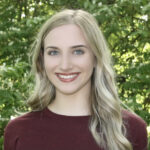 Katie Rodriguez headshot, wearing burgundy shirt, standing in front of greenery.