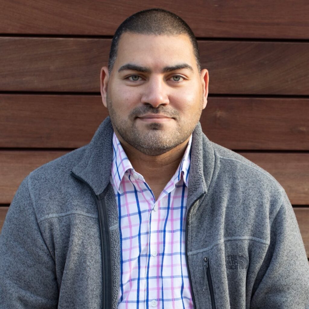 Raul Fernandez headshot, in gray fleece with checkered dress shirt, standing in front of brown wall.