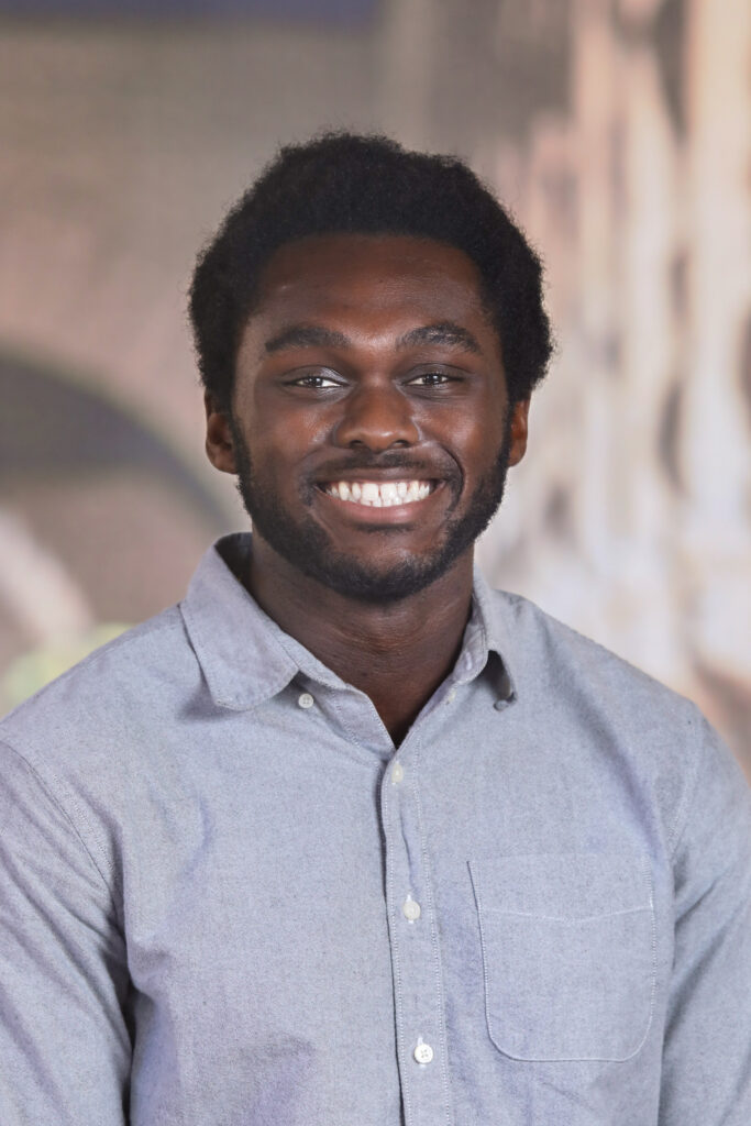 Second version of Cordy McJunkins headshot, gray shirt standing in front of white campus buildings.
