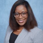 Tia Patrick headshot, wearing gray blazer and black shirt, standing in front of light blue background.