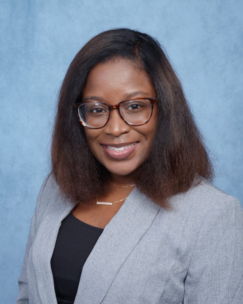 Tia Patrick headshot, wearing gray blazer and black shirt, standing in front of light blue background.