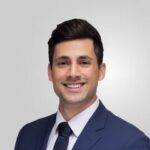 Kia Azadbakht headshot, navy blue suit, dark tie, white shirt, standing in front of white background.