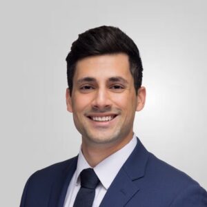 Kia Azadbakht headshot, navy blue suit, dark tie, white shirt, standing in front of white background.